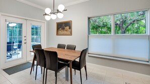 Et spiseområde med et træbord og sorte stole, en loftlampe, et billede på væggen og et vindue med persienner.