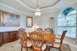 Une salle à manger avec une table et des chaises en bois, un lustre, un grand tableau et une fenêtre avec des stores.