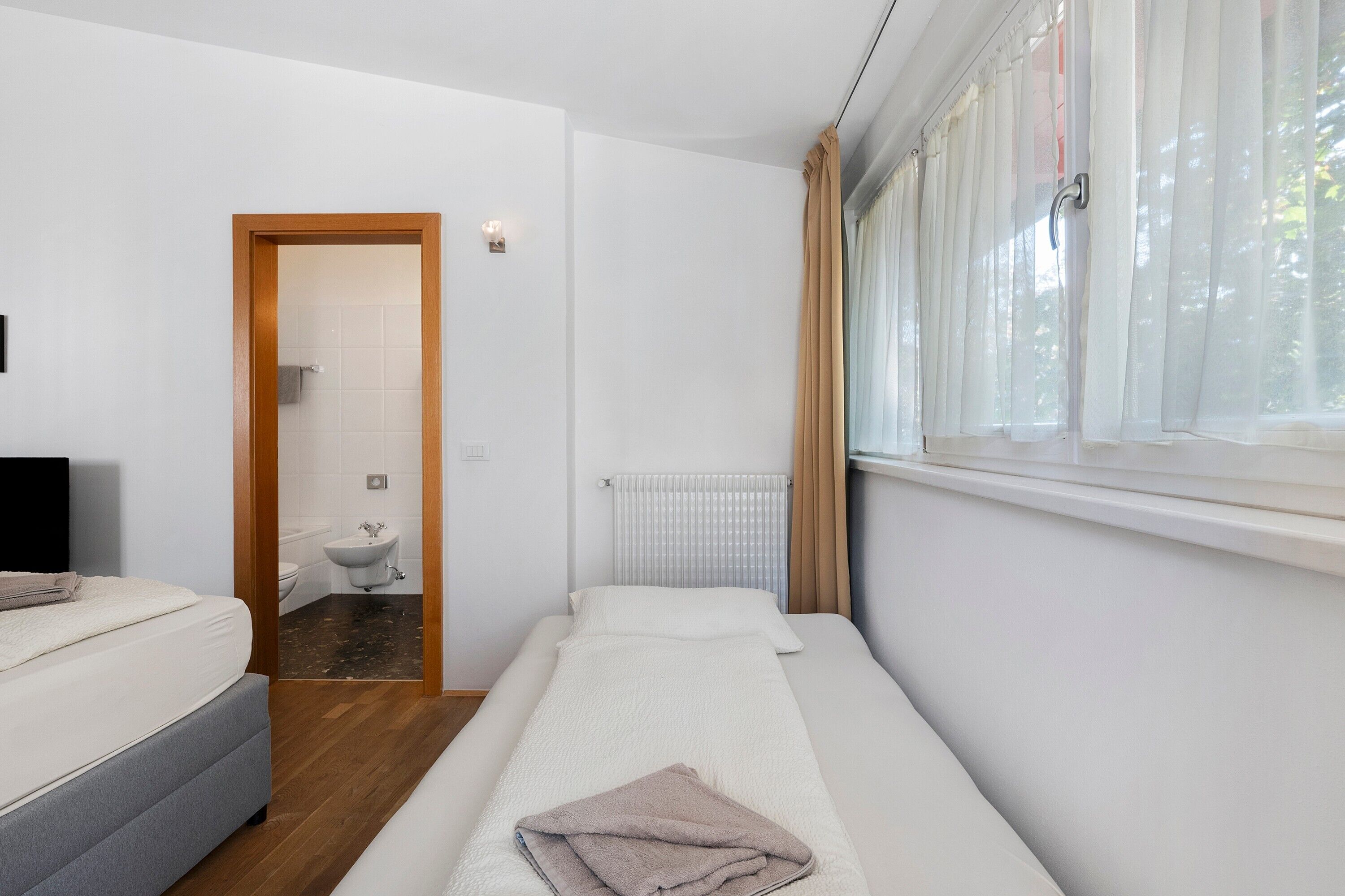 Una piccola sistemazione con una singola stanza, un letto, un comodino, un termosifone, una finestra con tende e un bagno con il water e un lavandino.