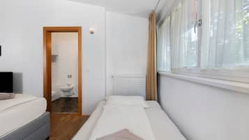Una piccola sistemazione con una singola stanza, un letto, un comodino, un termosifone, una finestra con tende e un bagno con il water e un lavandino.