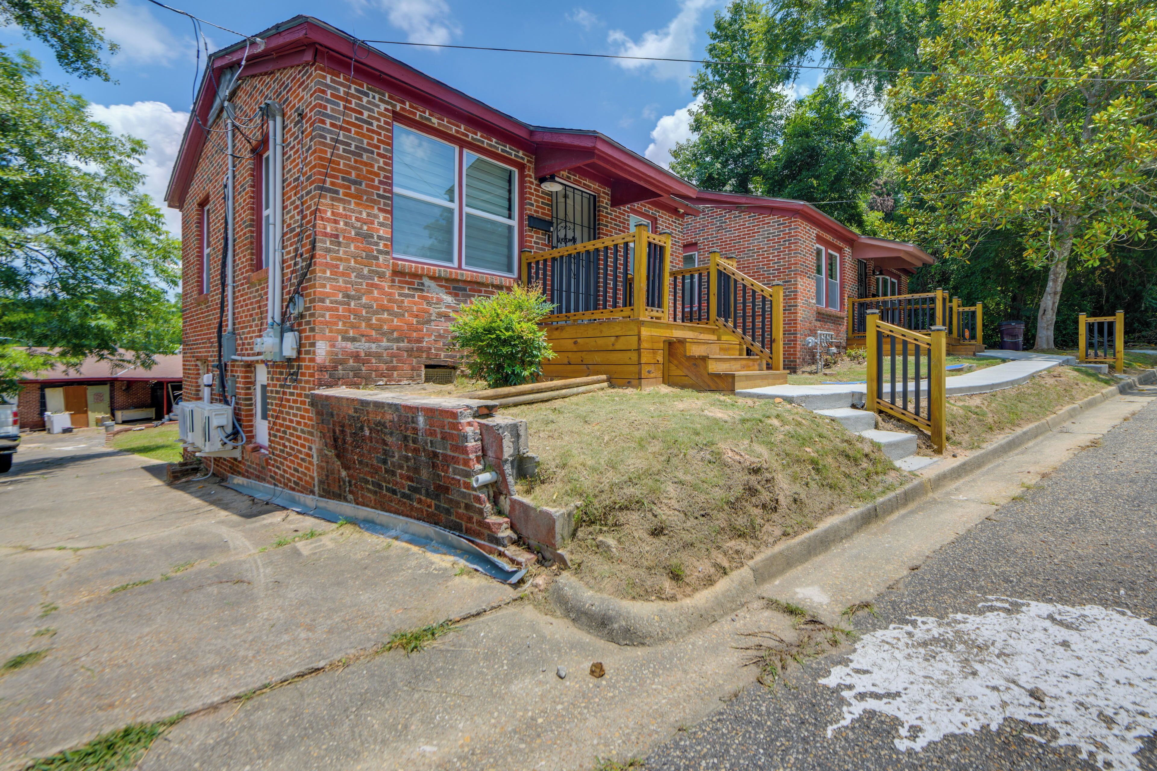 A two-story brick house with a red roof, a front porch with a wooden staircase, and a small front yard.