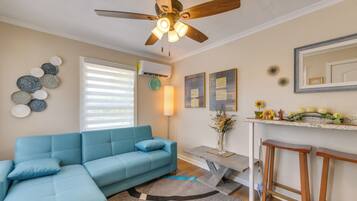 A living room with a blue sectional sofa, a ceiling fan, a wall-mounted air conditioner, a clock, decorative wall art, a vase with flowers, and a kitchen counter with fruit and a mirror.