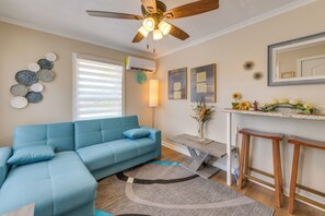 A living room with a blue sectional sofa, a ceiling fan, a wall-mounted air conditioner, a clock, decorative wall art, a vase with flowers, and a kitchen counter with fruit and a mirror.