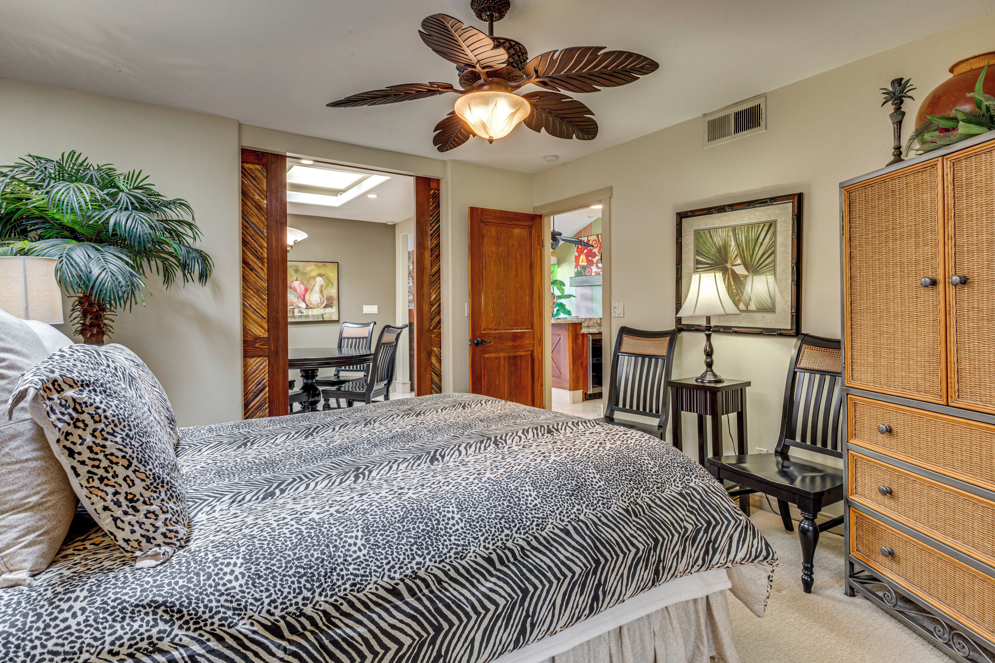 A bedroom with a zebra-patterned bedspread, a ceiling fan, a large wooden wardrobe, and a dining area visible through an open door.