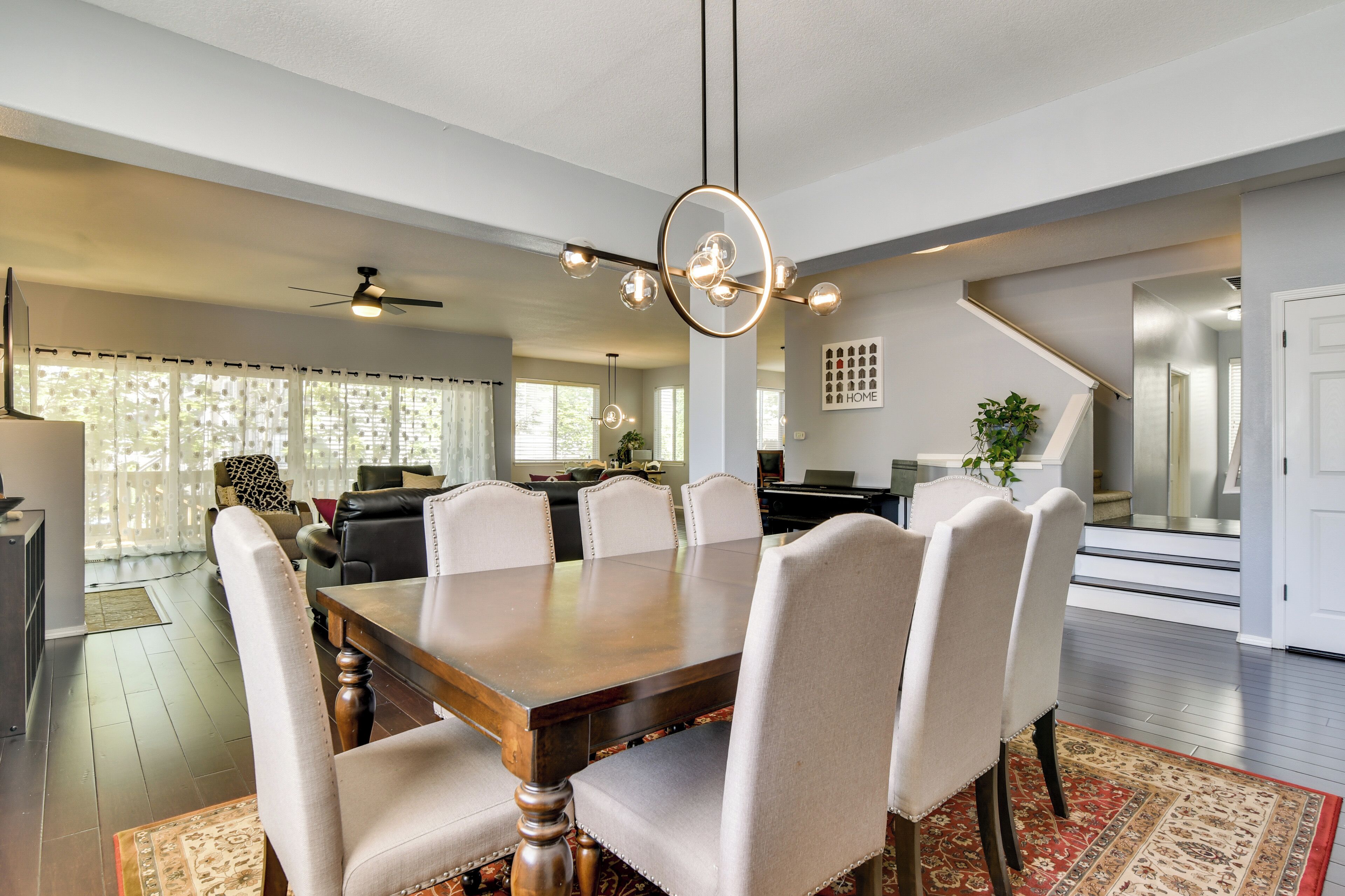 Un coin repas avec une table en bois et des chaises rembourrées, un ventilateur de plafond et, au fond, une cuisine.