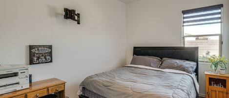 A bedroom with a bed, a desk with a printer, a wall-mounted shelf, and a window with blinds.