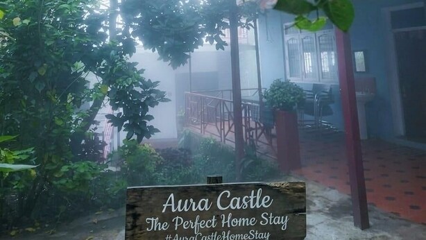 Een binnenplaats met een gebouw met rode dakpannen, omringd door groen en een bordje met de tekst 'Aura Castle The Perfect Home Stay'.