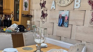 A dining area with a wooden table set for four, surrounded by upholstered chairs with rivets. A large wall clock and decorative elements are visible above the table. - BridAj's Place is a full Semi-detached 3 Bed home with parking full of History. (Belfast)