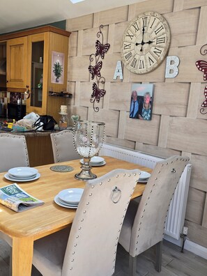 A dining area with a wooden table set for four, surrounded by upholstered chairs with rivets. A large wall clock and decorative elements are visible above the table. - BridAj's Place is a full Semi-detached 3 Bed home with parking full of History. (Belfast)