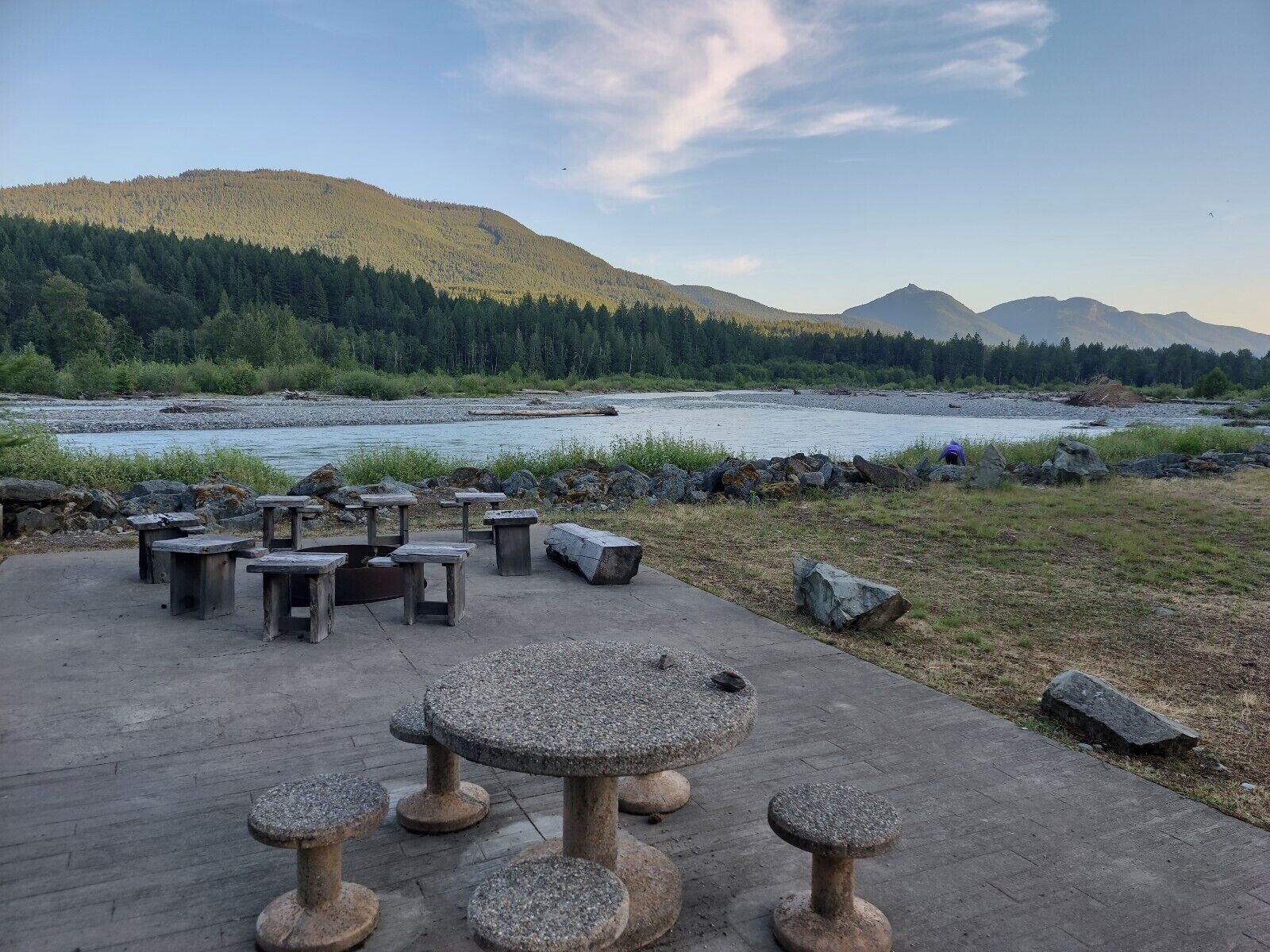 Een terras met stenen tafels en banken, met uitzicht op een rivier en beboste heuvels.