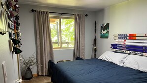 Une chambre avec un lit, une fenêtre avec des rideaux, un porte-skis fixé au mur et un tableau encadré.