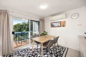 A room with a dining table, chairs, a potted plant, a clock, and an air conditioning unit. - Charming 2-Bed Coastal Haven Near Esplanade (Maroochydore)