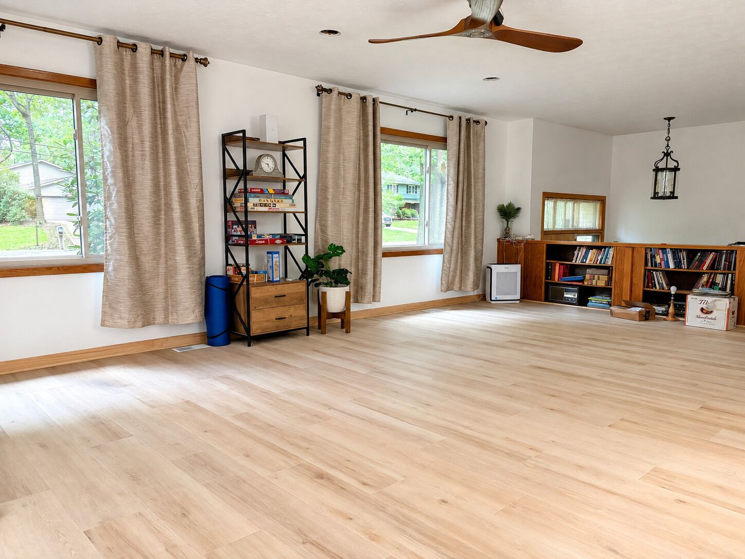 Una habitación amplia con ventanas grandes, un ventilador de techo, una estantería y un pequeño escritorio con una planta.