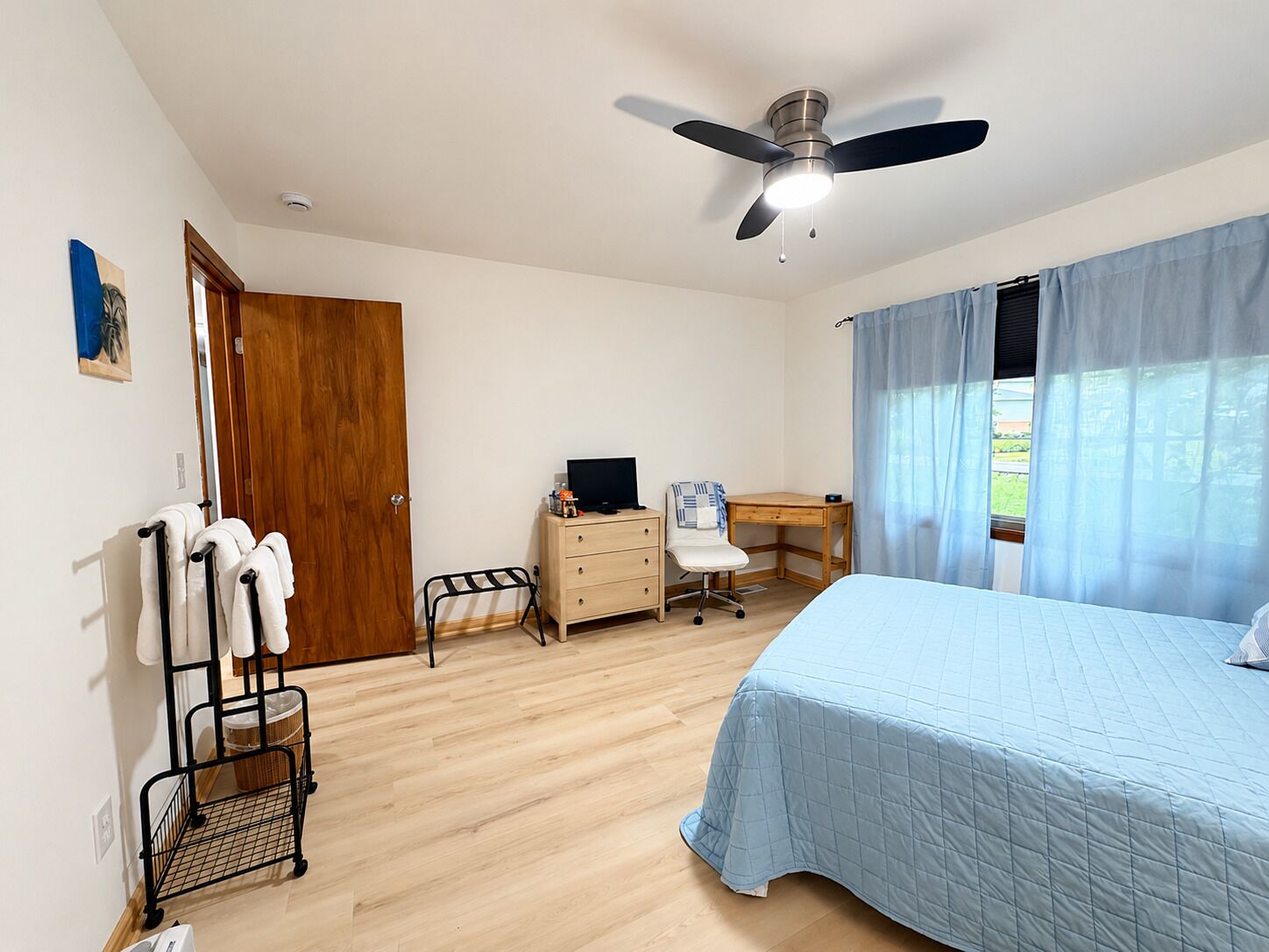 A bedroom with a bed, a desk, a chair, a ceiling fan, and a window with curtains.