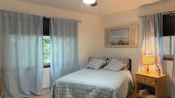 A bedroom with a bed, curtains, a ceiling fan, a bedside table with a lamp, and a framed picture on the wall.