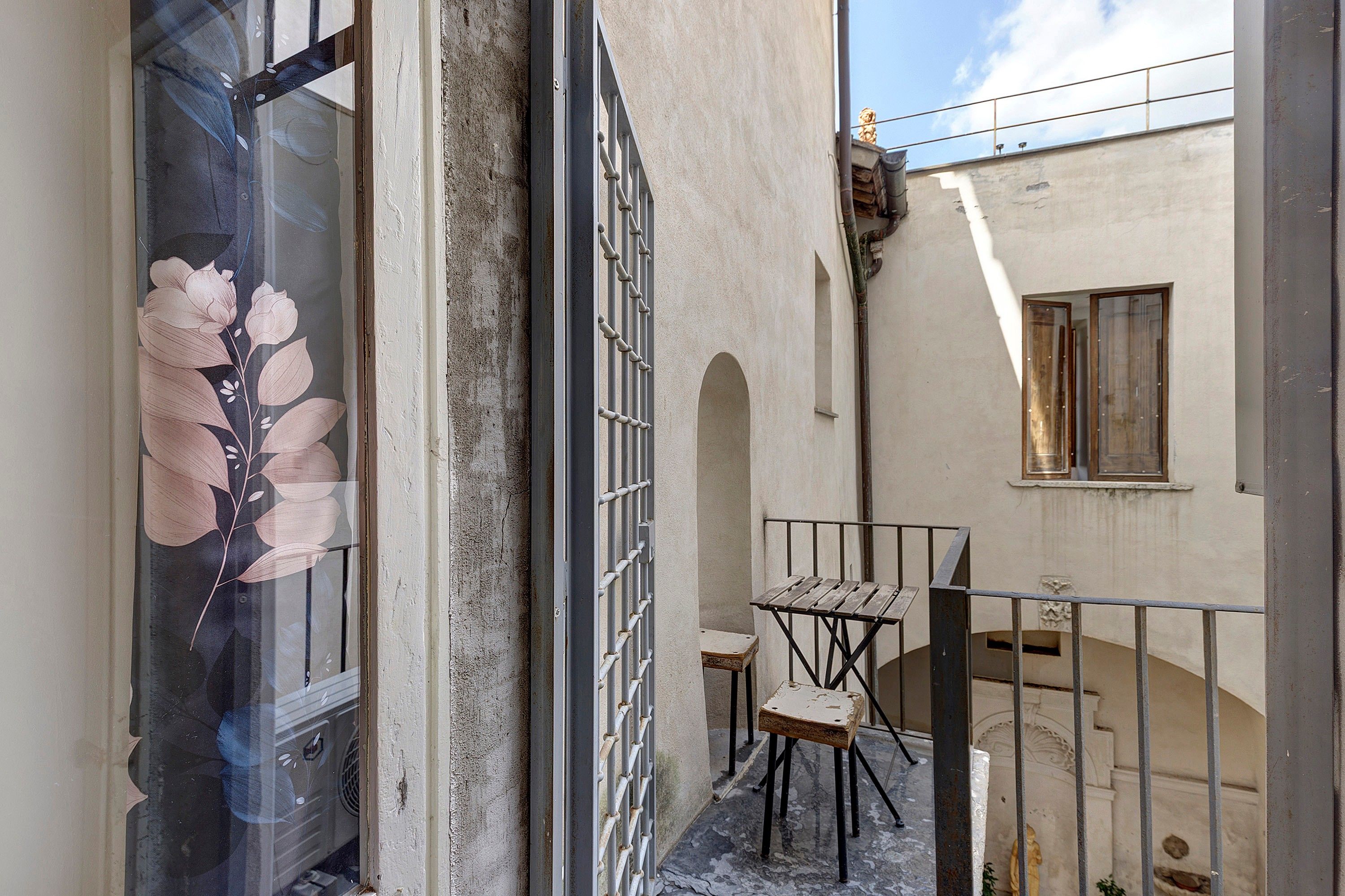 A narrow outdoor corridor with a metal gate, a small table with chairs, and a window with a floral design.