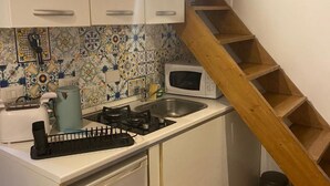 Eine kleine Küche mit weißen Schränken, einem Gasherd, einer Mikrowelle und einem Spülbecken. Eine Holztreppe befindet sich rechts.