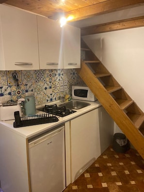 A compact kitchen with white cabinets, a gas stove, a microwave, and a sink. There's a wooden staircase to the right. - Little House in Trastevere Apartment with Balcony, Wi-Fi and Air Conditioning (Rome)