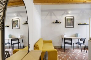 Ein Zimmer mit einem gelben Sofa, zwei schwarzen Stühlen, einem weißen Tisch mit Wasserkocher und zwei Regalen mit Dekoration.
