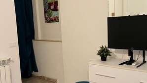 Una habitación con una cama, un escritorio con computadora, una silla, una planta pequeña y un cuadro en la pared.