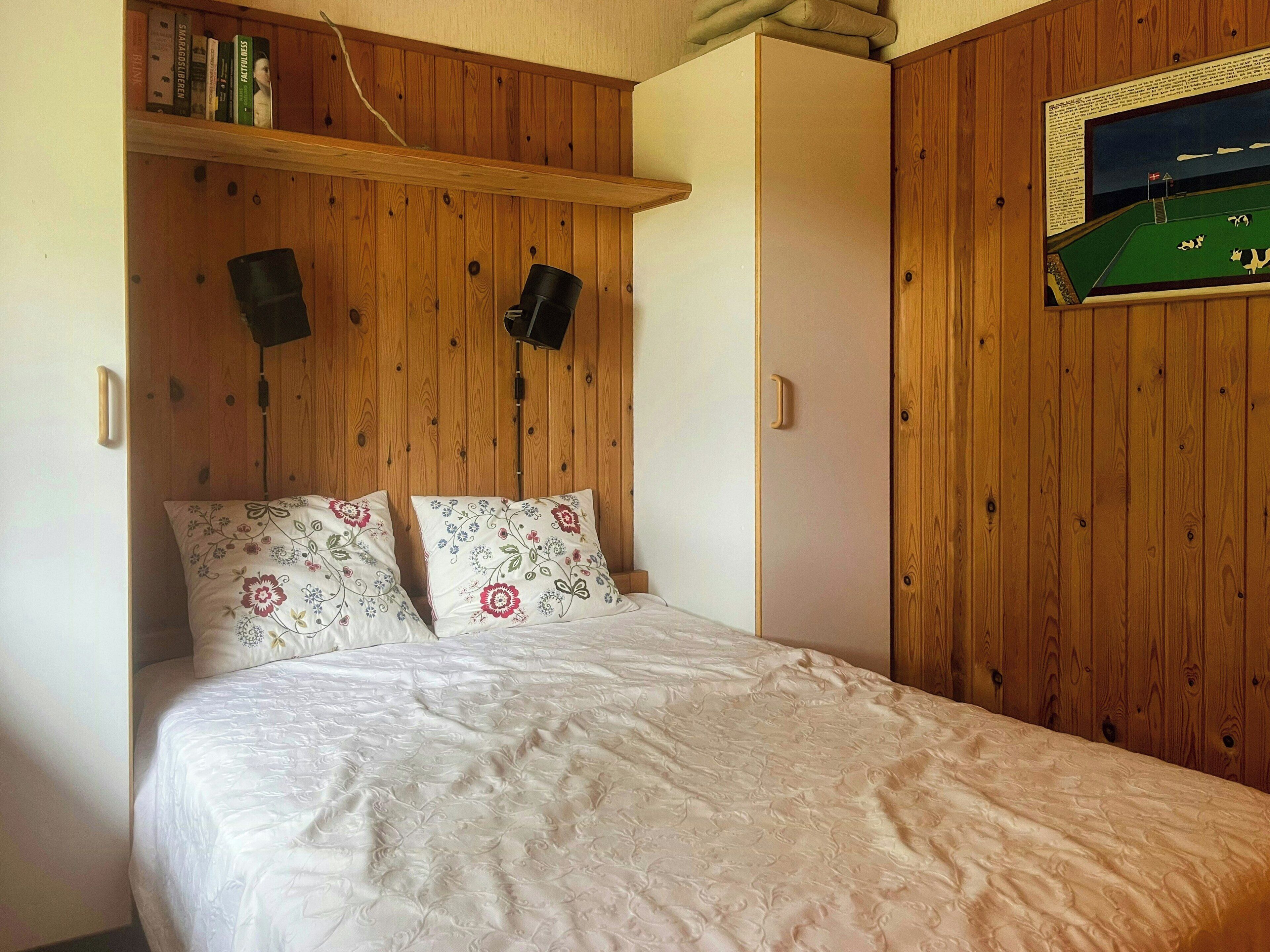 A bedroom with a wooden headboard, a bed with floral-patterned pillows, a nightstand, and a wall-mounted shelf with books.