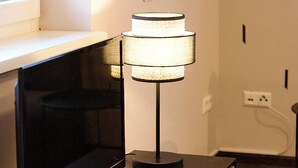 Een televisie op een houten standaard, een lamp met een structuurrijke kap, en een muur met verticale jaloezieën en ingelijste foto's.