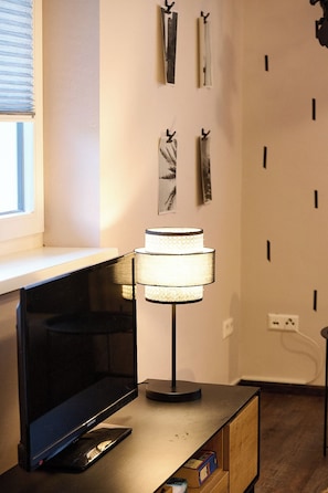 Ein Fernseher auf einem Holzständer, eine Lampe mit einer strukturierten Schirm und eine Wand mit vertikalen Jalousien und gerahmten Bildern.