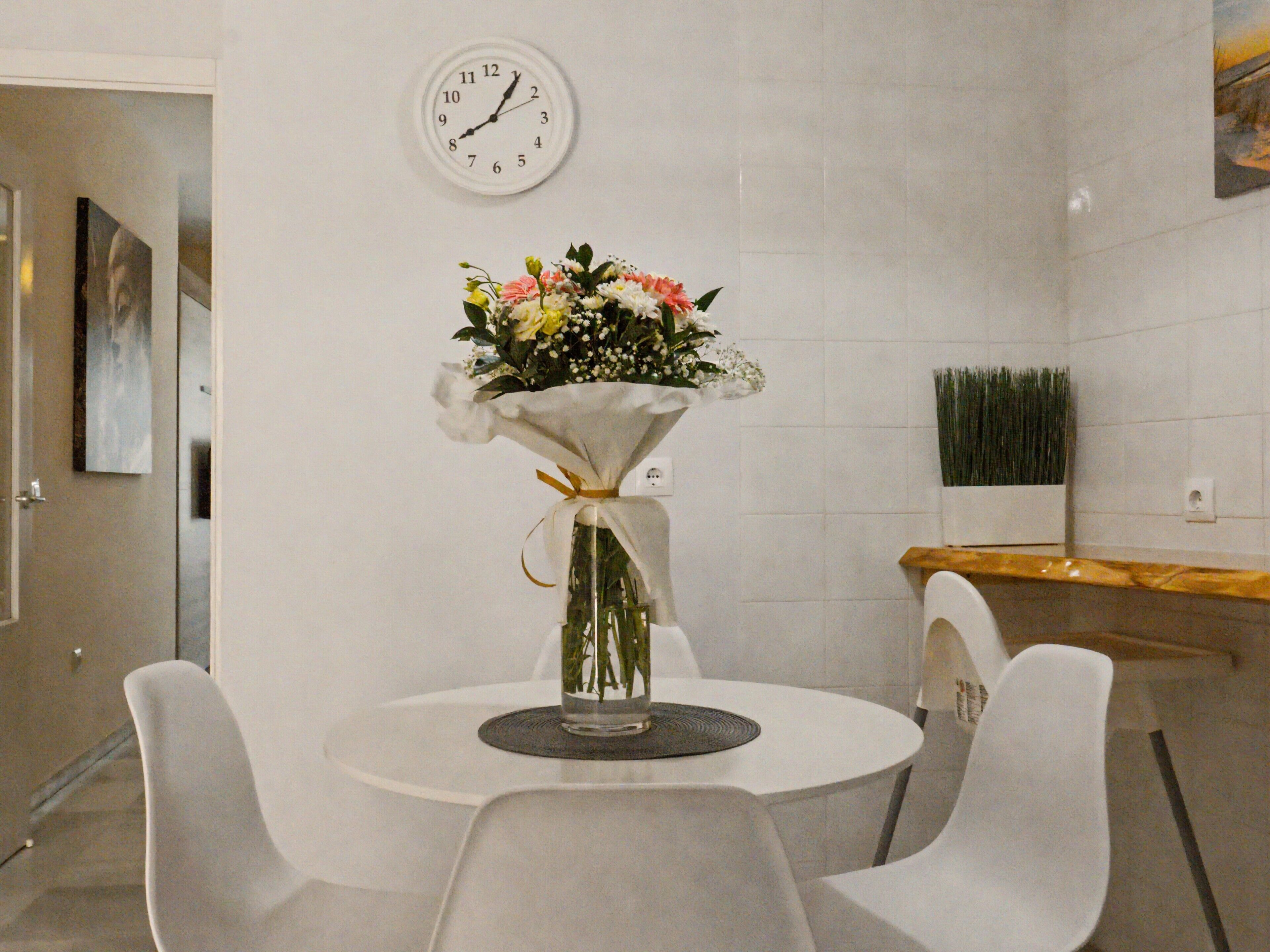 Un coin repas avec une table ronde, des chaises blanches, une horloge au mur et un arrangement floral sur la table.