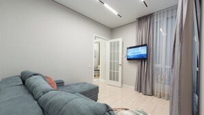A modern living room with a grey sofa, a flat-screen TV mounted on the wall, and a door leading to another room. - Spacious 3-room apartments (Almaty)