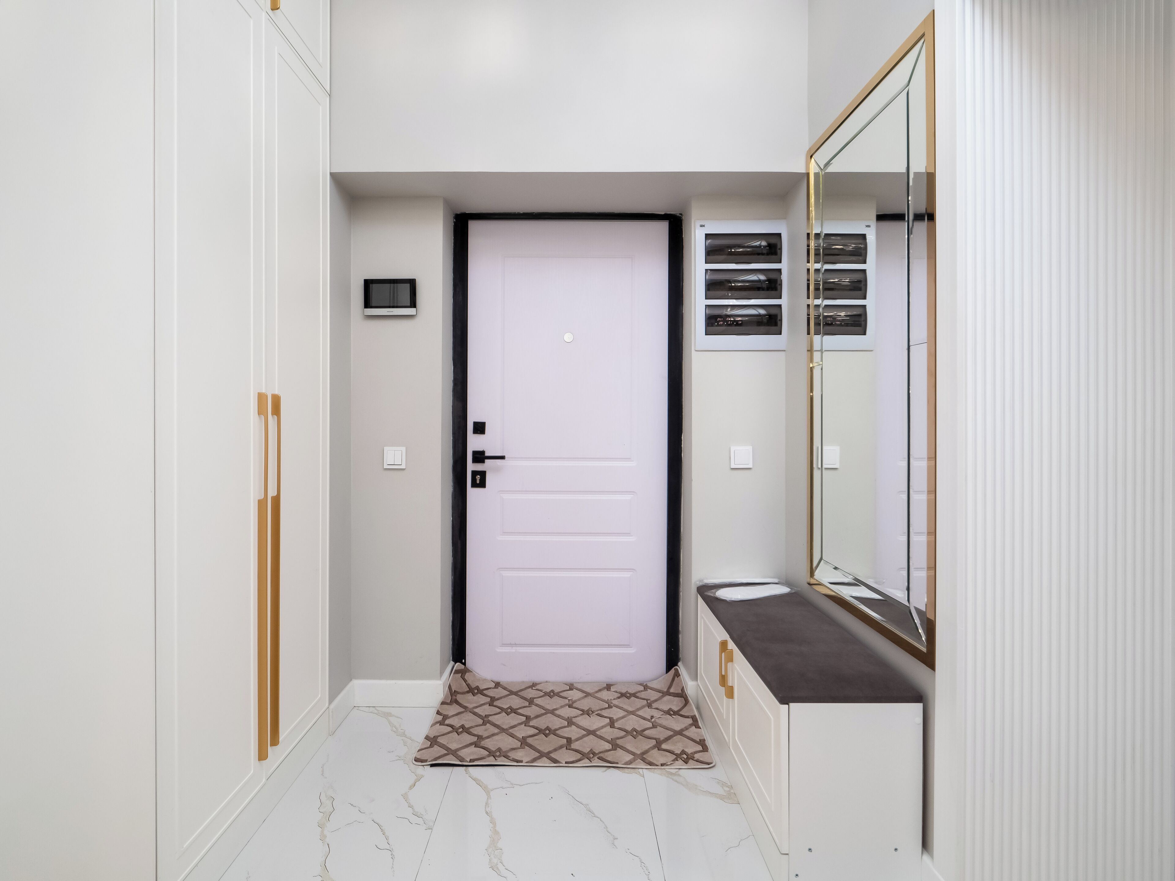 Un couloir avec une porte blanche, un petit tapis, un miroir et une fenêtre à cadre blanc.