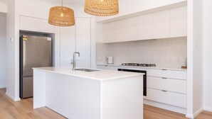 A modern kitchen with a central island, stainless steel appliances, and white cabinetry. - Neptune Coastal Retreat (Forster)