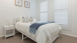 A bedroom with a bed, a nightstand, a lamp, and two framed pictures on the wall. - Neptune Coastal Retreat (Forster)