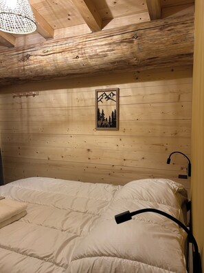 Teto de madeira com um quadro na parede, uma cama com roupa de cama branca e um abajur preto.