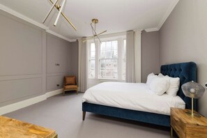 Een slaapkamer met een groot bed, een stoel, een houten nachtkastje en een raam met gordijnen.