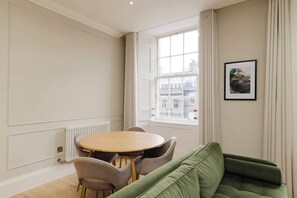 A room with a round table and chairs, a green sofa, a radiator, and a window with a view of buildings. - Stylish Apartment on Castle Street (Edinburgh)