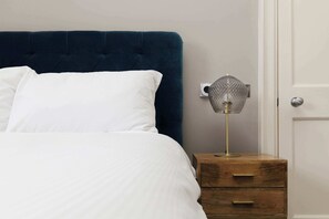A bed with white bedding, a dark blue headboard, a bedside table with a lamp, and a wooden nightstand. - Stylish Apartment on Castle Street (Edinburgh)