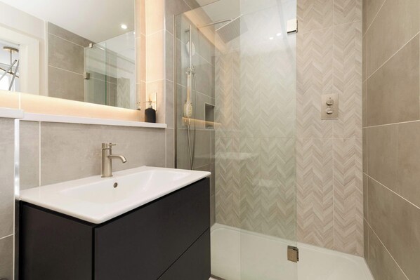 A modern bathroom with a white sink, dark cabinet, glass shower enclosure, and herringbone patterned tiles. - Stylish Apartment on Castle Street (Edinburgh)