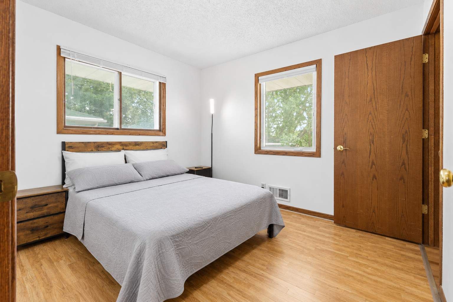 Une chambre à coucher comprenant un lit, des tables de chevet, une armoire et deux fenêtres.