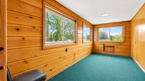 Une pièce aux murs en bois, avec une moquette verte et deux fenêtres donnant sur des arbres.