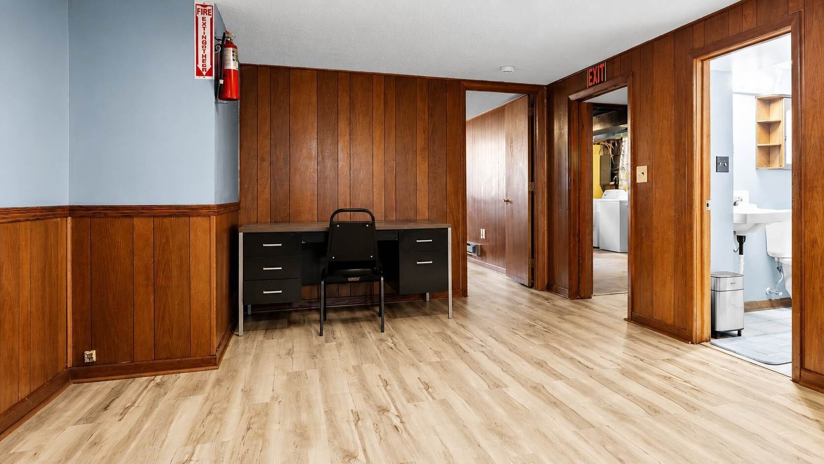 Una habitación con un escritorio de madera, una silla, un extintor y un letrero de salida.