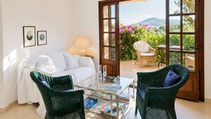 Ein Zimmer mit einem weißen Sofa, zwei grünen Korbstühlen, einem Glastisch mit Zeitschriften, einer Lampe und einer Tür, die zu einem Garten mit rosa Blumen und Blick auf Hügel führt.