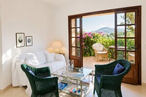 Ein Zimmer mit einem weißen Sofa, zwei grünen Korbstühlen, einem Glastisch mit Zeitschriften, einer Lampe und einer Tür, die zu einem Garten mit rosa Blumen und Blick auf Hügel führt.