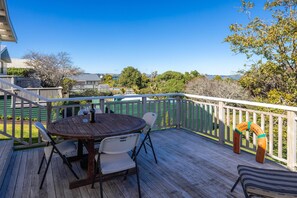 A wooden deck with a round table and chairs, a lifebuoy, and a view of a residential area with houses and trees. - Beautifully renovated three-bedroom holiday home sleeping 4 adults and 2 kids (Waiheke Island)