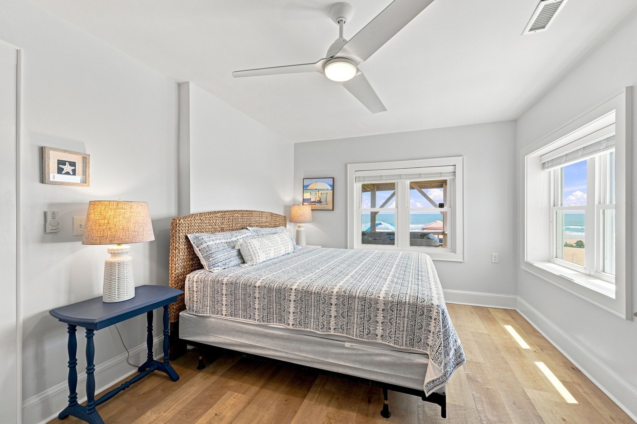 Une chambre à coucher comprenant un lit, une table de chevet, une lampe, un ventilateur de plafond, une fenêtre avec vue et un tableau accroché au mur.