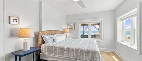A bedroom with a bed, a nightstand, a lamp, a ceiling fan, a window with a view, and a wall-mounted picture.