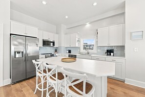 A modern kitchen with white cabinetry, stainless steel appliances, and a central island with seating. - Luxury Oceanfront Home w/ Hot tub and Community Pool (North Topsail Beach)