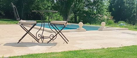 Un espace aménagé au bord de la piscine, avec une table et des chaises en métal, entouré de pelouse et d’arbres.
