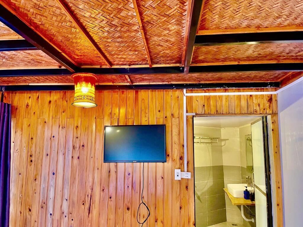 Ett rum med träpanel på väggarna, en tv som hänger på väggen, en duschhörna som syns genom en glasdörr och ett vävt tak.