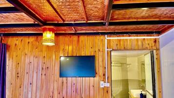 A wooden-paneled room with a television mounted on the wall, a shower area visible through a glass door, and a woven ceiling.