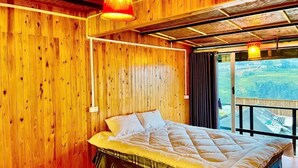 Una habitación de madera con una cama, un ventanal con vistas y una lámpara colgante.